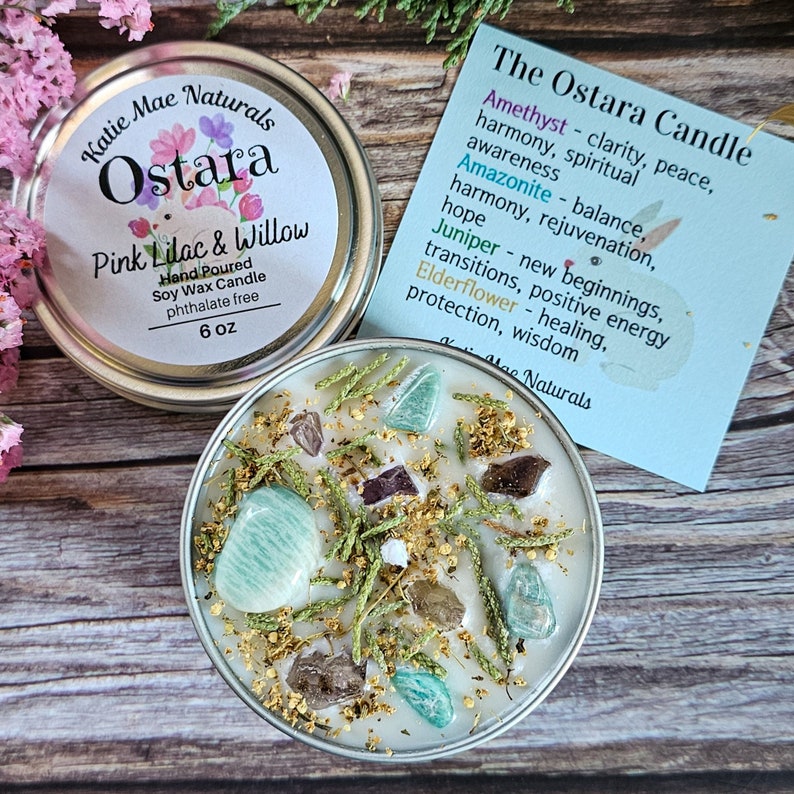 Peut inclure: Une bougie en cire de soja blanche dans un r&eacute;cipient en &eacute;tain avec un couvercle. La bougie est d&eacute;cor&eacute;e de brindilles vertes, de petits cristaux et de paillettes dor&eacute;es. L'&eacute;tiquette sur le couvercle indique "Ostara Pink Lilac & Willow Hand Poured Soy Wax Candle phthalate free 6 oz". Une carte s&eacute;par&eacute;e indique "The Ostara Candle Amethyst - clart&eacute;, paix, harmonie, conscience spirituelle Amazonite - &eacute;quilibre, harmonie, rajeunissement, espoir Gen&eacute;vrier - nouveaux commencements, transitions, &eacute;nergie positive Sureau - gu&eacute;rison, protection, sagesse Katie Mae Naturals".