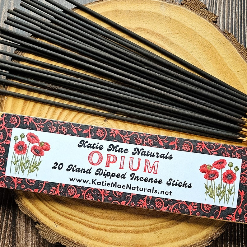 May include: Black incense sticks with gold tips are arranged on a wooden surface. A box of "Opium" incense sticks is visible, with the text "Katie Mae Naturals" and "20 Hand Dipped Incense Sticks". The box features red poppy illustrations and a decorative border.