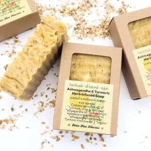 May include: A bar of handmade, all-natural, vegan Ashwagandha & Turmeric Herb Infused Soap, scented with midseason orange. The soap is packaged in a brown cardboard box with a white label that lists the ingredients: coconut, olive, sunflower, ashwagandha oil, kokum butter, midseason orange essential oil, ashwagandha root powder, turmeric root powder, and organic. The label also includes the brand name "Katie Mae Naturals".
