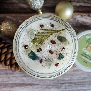 Puede incluir: Una vela de cera de soja en un tarro de vidrio transparente, decorada con piedras preciosas, purpurina y una ramita de vegetaci&oacute;n. La vela est&aacute; rodeada de adornos navide&ntilde;os y una pi&ntilde;a. La etiqueta de la tapa dice "Cozy Cabin".