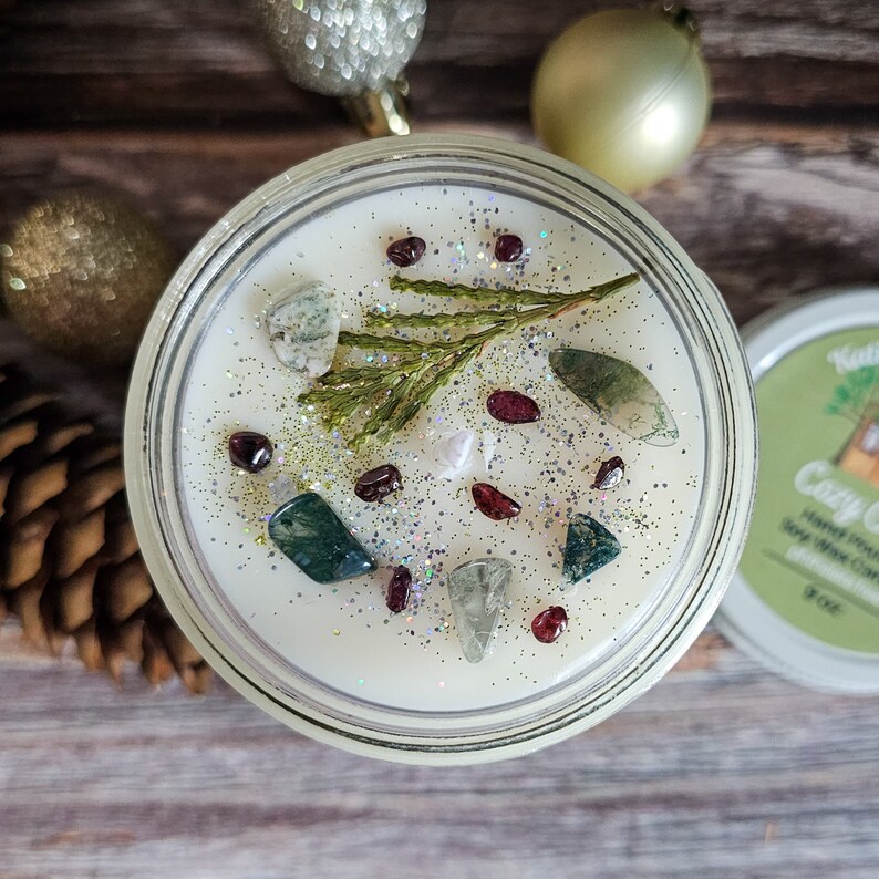 Puede incluir: Una vela de cera de soja en un tarro de vidrio transparente, decorada con piedras preciosas, purpurina y una ramita de vegetaci&oacute;n. La vela est&aacute; rodeada de adornos navide&ntilde;os y una pi&ntilde;a. La etiqueta de la tapa dice "Cozy Cabin".