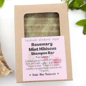 Może przedstawiać: Brązowe pudełko kartonowe z r&oacute;żowo-białą etykietą z napisem "Rosemary Mint Hibiscus Shampoo Bar" i "4 oz (approx.)". Etykieta zawiera listę składnik&oacute;w, w tym kokos, oliwę z oliwek, słonecznik, olej rycynowy, pestki winogron, masło kakaowe, proszek z hibiskusa, rozmaryn i olejki eteryczne z mięty pieprzowej. Etykieta informuje r&oacute;wnież, że produkt jest ręcznie robiony, w 100% naturalny, wegański i organiczny. Etykieta pochodzi od Katie Mae Naturals.