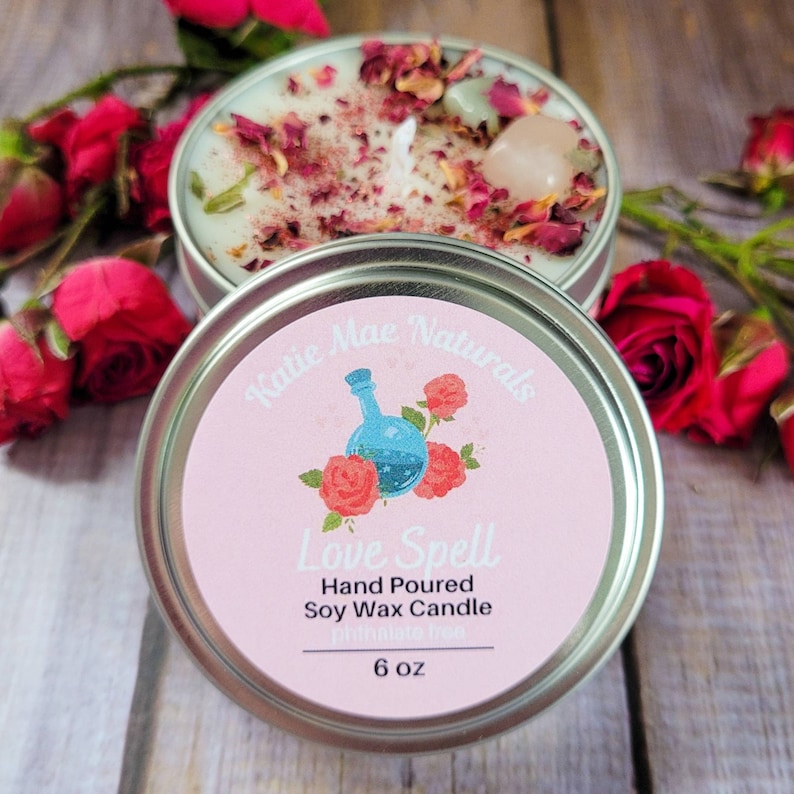 May include: A pink and silver candle tin with a label that reads "Katie Mae Naturals Love Spell Hand Poured Soy Wax Candle 6 oz". The label features a blue potion bottle with roses and a pink background. The candle tin is filled with white wax and rose petals.