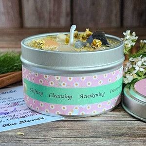 May include: A silver tin candle with a white wick and various crystals and dried flowers on top. The label on the tin reads "Shifting Cleansing Awakening Introspection".