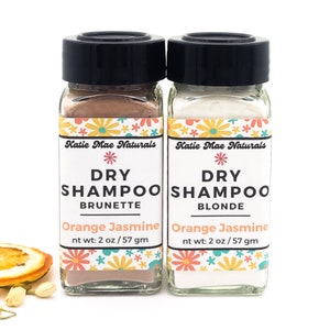 May include: Two small glass jars with black lids containing dry shampoo powder. The jar on the left is labeled "Dry Shampoo Brunette Orange Jasmine" and the jar on the right is labeled "Dry Shampoo Blonde Orange Jasmine". Both jars have a floral design on the label.