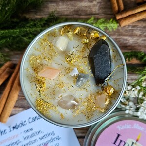 May include: A white candle with gold glitter and various crystals, including a large black crystal, in a silver tin. The candle is labeled "The Imbolc Candle" and has text about the candle's purpose.
