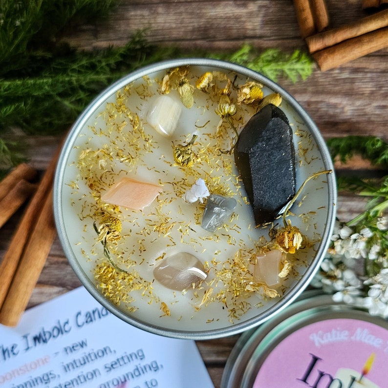 May include: A white candle with gold glitter and various crystals, including a large black crystal, in a silver tin. The candle is labeled "The Imbolc Candle" and has text about the candle's purpose.