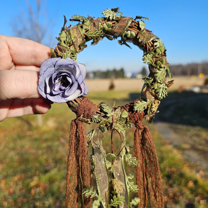 May include: A purple flower with brown and green leaves attached to a brown and green wreath with brown and green tassels hanging from the bottom.