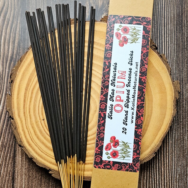 May include: A bundle of black incense sticks with gold tips, next to a kraft paper box. The box features the text "Opium" and "20 Hand Dipped Incense Sticks" with a floral design and the brand name "Katie Mae Naturals".