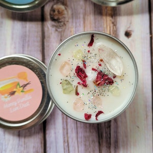 Puede incluir: Una vela blanca con acentos de cristal rosa claro, amarillo y transparente, p&eacute;talos de rosa y purpurina plateada. La vela est&aacute; en una lata de plata con el texto "Honey Vanilla Love Dust" en la etiqueta.