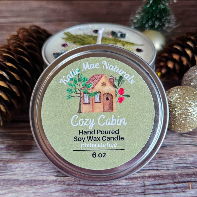 May include: A silver tin candle with a green label that says "Katie Mae Naturals Cozy Cabin Hand Poured Soy Wax Candle phthalate free 6 oz". The label also features a cartoon illustration of a small house with a tree and red berries.