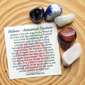 May include: A card with the text "Mabon - Autumnal Equinox" and a description of the holiday. The card is surrounded by six colorful stones, including black, blue, white, red, and pink.