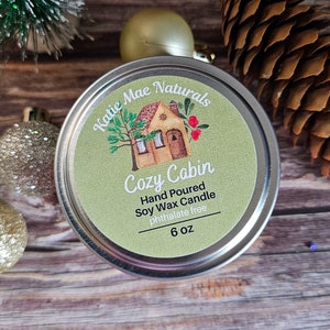 May include: A 6 ounce soy wax candle in a silver tin with a green label. The label features a cartoon illustration of a small house with a red roof and a tree. The text on the label reads "Katie Mae Naturals Cozy Cabin Hand Poured Soy Wax Candle Phthalate Free 6 oz".