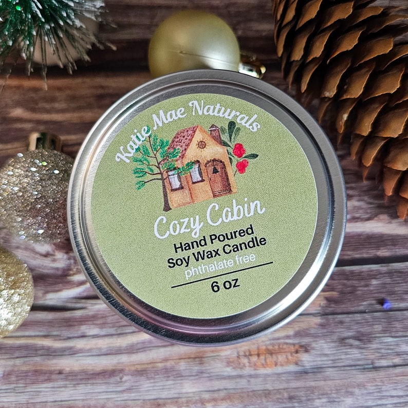 May include: A 6 ounce soy wax candle in a silver tin with a green label. The label features a cartoon illustration of a small house with a red roof and a tree. The text on the label reads "Katie Mae Naturals Cozy Cabin Hand Poured Soy Wax Candle Phthalate Free 6 oz".