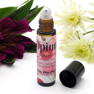 Puede incluir: Un peque&ntilde;o frasco de vidrio con una tapa de rodillo negra, lleno de un l&iacute;quido transparente. El frasco tiene una etiqueta rosa y blanca con el texto "Opium Perfume Oil 10 ml/0.4 oz contains: Sunflower Opium Fragrance Katie Mae Natur www.KatieMaeNaturals.com".