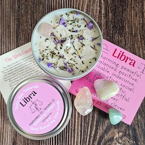 May include: A pink and white candle jar with a Libra zodiac symbol and the text "Katie Mae Naturals Libra" on the label. The candle is decorated with dried flowers, herbs, and small pink and white crystals. The candle is labeled "September 23 - October 22 Blueberry Cobbler Candle 6 oz Phthalate Free Hand Poured Soy Wax Candle".