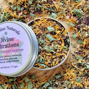 May include: A silver tin with a purple label that reads "Mae Naturals Divine Vibrations Herbal Incense" and lists the ingredients: lemon verbena, calendula, yarrow, hibiscus, and elderflower. The tin is open and filled with a mixture of dried herbs and flowers, including yellow calendula petals, green leaves, and small white flowers. The tin is sitting on a wooden surface surrounded by loose herbs and flowers.