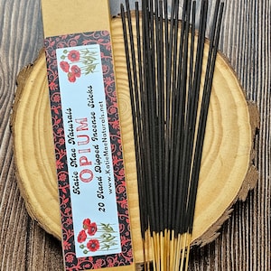 May include: A bundle of black incense sticks with light brown tips, next to a cardboard box with the text "Opium" and "20 Hand Dipped Incense Sticks". The box has a floral design with red poppies.
