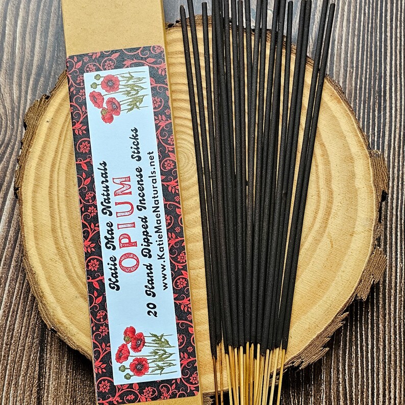 May include: A bundle of black incense sticks with light brown tips, next to a cardboard box with the text "Opium" and "20 Hand Dipped Incense Sticks". The box has a floral design with red poppies.