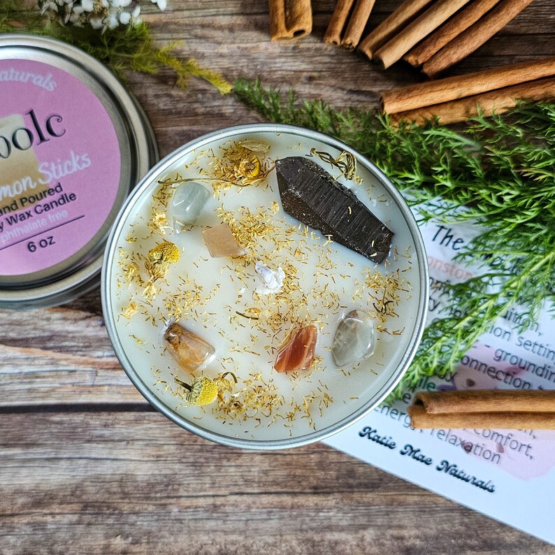 May include: A white soy wax candle with gold glitter, crystals, and dried flowers. The candle is in a silver tin with a label that reads "The Moonstone Collection" and "Katie Mae Naturals".