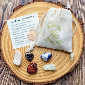 May include: A set of six tumbled gemstones in a small white drawstring bag with a green logo that says "Katie me Naturals". The stones are feldspar, hematite, sodalite, red tiger's eye, aquamarine, and a sixth stone that is not labeled. The stones are arranged on a wooden surface. The stones are for self-awareness, creativity, abundance, harmonizing mind-body-spirit, removing limitations, intuition, clarity, communication, balance, strength, confidence, courage, tolerance, generosity, and overcoming obstacles.