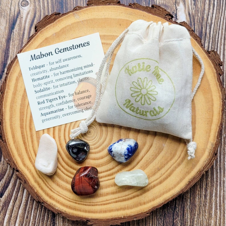 May include: A set of six tumbled gemstones in a small white drawstring bag with a green logo that says "Katie me Naturals". The stones are feldspar, hematite, sodalite, red tiger's eye, aquamarine, and a sixth stone that is not labeled. The stones are arranged on a wooden surface. The stones are for self-awareness, creativity, abundance, harmonizing mind-body-spirit, removing limitations, intuition, clarity, communication, balance, strength, confidence, courage, tolerance, generosity, and overcoming obstacles.