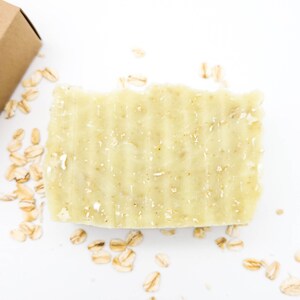 May include: A light green bar of soap with oatmeal flakes embedded in it. The soap is on a white surface with scattered oatmeal flakes.