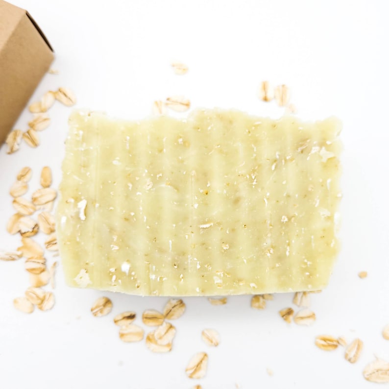 May include: A light green bar of soap with oatmeal flakes embedded in it. The soap is on a white surface with scattered oatmeal flakes.