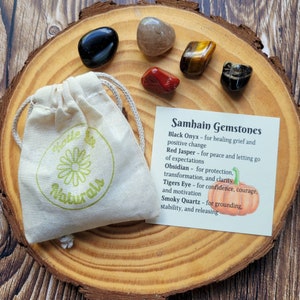 May include: A set of six polished gemstones in a small white drawstring bag, with a card listing the stones and their properties. The stones are black onyx, red jasper, clear quartz, tiger's eye, obsidian, and smoky quartz. The card reads "Samhain Gemstones" and lists the properties of each stone.