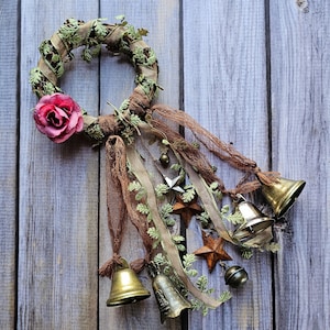 May include: A rustic wreath made of brown twigs and green leaves with a pink fabric rose, decorated with bells, stars, and ribbons.