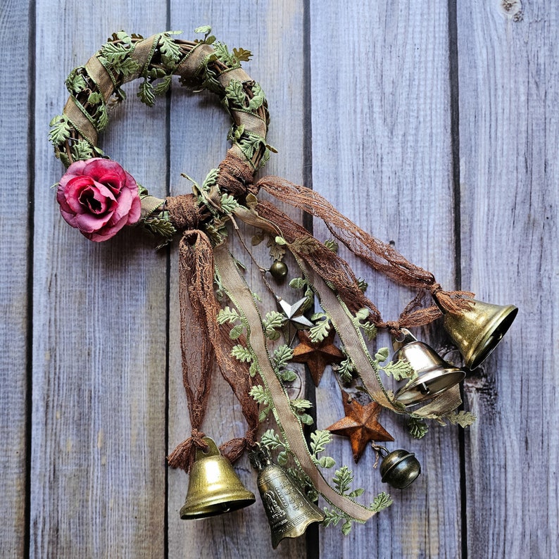 May include: A rustic wreath made of brown twigs and green leaves with a pink fabric rose, decorated with bells, stars, and ribbons.