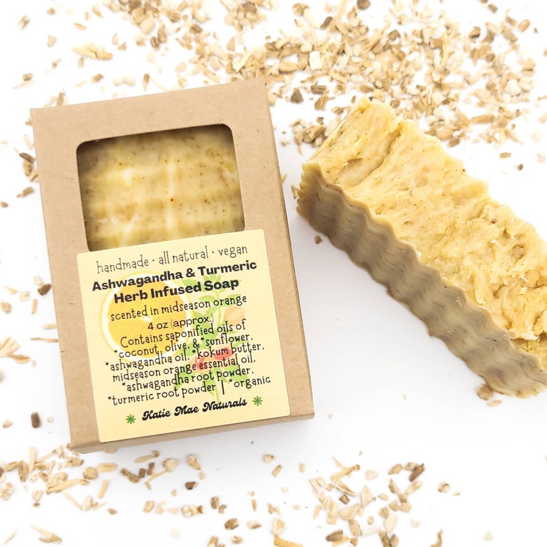 May include: A 4 ounce bar of handmade, vegan, Ashwagandha & Turmeric Herb Infused Soap, scented in midseason orange. The soap is in a cardboard box with a label that lists the ingredients: coconut, olive, sunflower, ashwagandha oil, kokum butter, midseason orange essential oil, ashwagandha root powder, turmeric root powder, and organic. The soap is sitting on a white surface with dried herbs scattered around it.