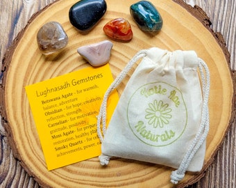 May include: A set of seven gemstones, including Botswana agate, obsidian, carnelian, moss agate, and smoky quartz, arranged on a wooden surface with a small cloth bag labeled "Dottie Mae Naturals". The gemstones are associated with various properties, such as warmth, balance, hope, creativity, strength, motivation, positivity, preparation, restoration, and power.