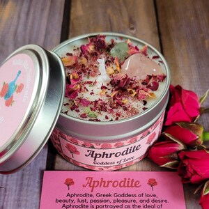 May include: A white candle with rose petals and crystals in a silver tin. The candle is labeled "Aphrodite Goddess of Love".