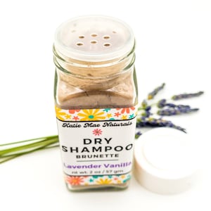 May include: A glass jar of dry shampoo for brunettes, labeled "Katie Mae Naturals Dry Shampoo Brunette Lavender Vanilla 2 oz / 57 gm". The jar has a clear lid with holes for dispensing the powder.