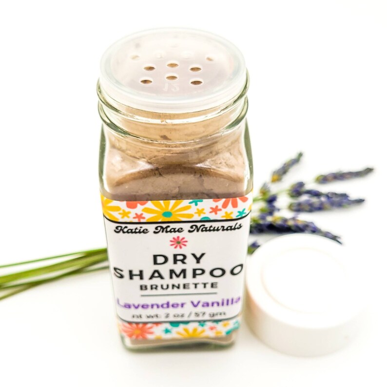May include: A glass jar of dry shampoo for brunettes, labeled "Katie Mae Naturals Dry Shampoo Brunette Lavender Vanilla 2 oz / 57 gm". The jar has a clear lid with holes for dispensing the powder.