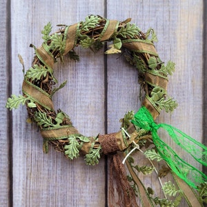 Pu&ograve; includere: Una ghirlanda verde e marrone con foglie verdi e nastro marrone. La ghirlanda &egrave; decorata con un nastro di pizzo verde e bianco.