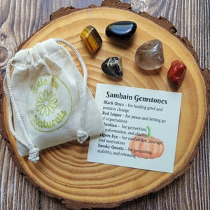 May include: A set of Samhain gemstones, including black onyx, red jasper, obsidian, tiger's eye, and smoky quartz, on a wooden surface with a small white bag and a card that reads "Samhain Gemstones" with descriptions of each stone's properties.