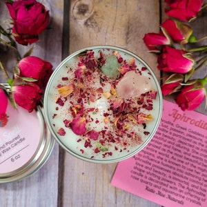 May include: A white candle with rose petals and crystals inside a glass jar. The candle is labeled "Spell" and "Hand Poured Wax Candle". A pink card with the text "Aphrodite" and a description of the Greek goddess of love, beauty, and pleasure is in the background.