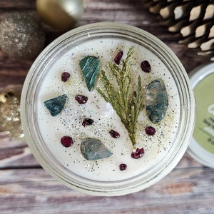 Puede incluir: Una vela de cera de soja en un tarro de cristal transparente, adornada con piedras decorativas, purpurina y una ramita de vegetaci&oacute;n. La superficie de la vela es blanca, y las piedras son verdes y rojas. Un art&iacute;culo decorativo para el hogar.