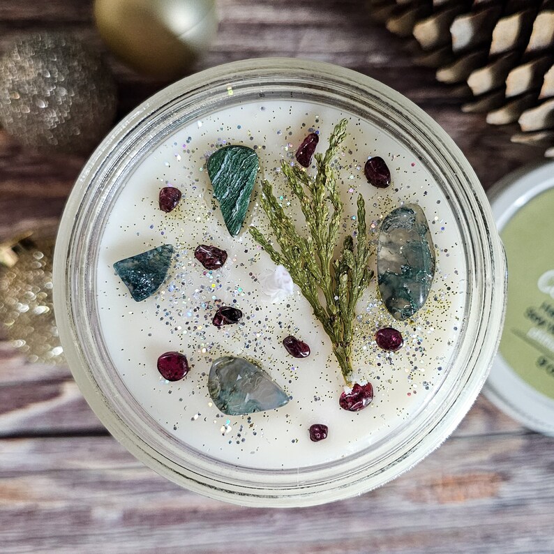 Puede incluir: Una vela de cera de soja en un tarro de cristal transparente, adornada con piedras decorativas, purpurina y una ramita de vegetaci&oacute;n. La superficie de la vela es blanca, y las piedras son verdes y rojas. Un art&iacute;culo decorativo para el hogar.