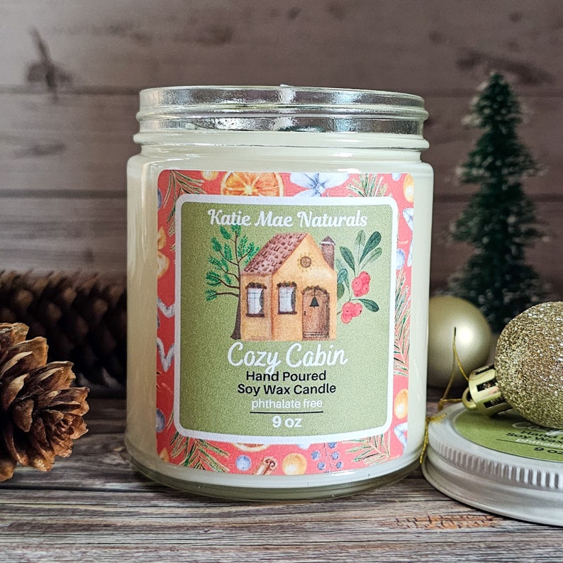 Puede incluir: Una vela en tarro de vidrio con una etiqueta que dice "Cozy Cabin". La etiqueta presenta un dibujo de una caba&ntilde;a, con las palabras "Hand Poured Soy Wax Candle" y "phthalate free". La vela es de 255 gramos.