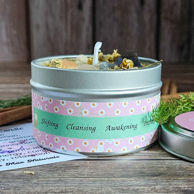 May include: A silver tin candle with a white wick, surrounded by crystals and dried flowers. The label on the tin reads "Shifting Cleansing Awakening Introsp".