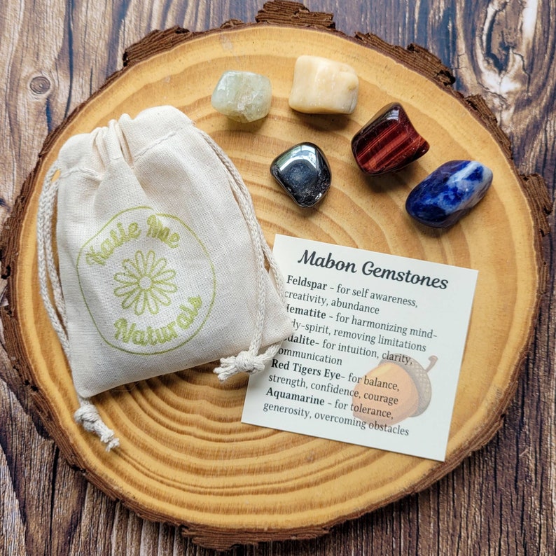 May include: A set of seven crystals on a wooden surface. The crystals are labeled with their names and properties. The crystals are: Feldspar, Hematite, Dalite, Red Tigers Eye, Aquamarine, and two other crystals. The crystals are arranged in a circle around a small white bag.