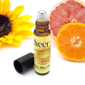 May include: A brown glass roller bottle with a silver roller ball. The bottle is labeled "cheer" and contains essential oils. The label also lists the ingredients: Sunflower, Grapefruit, and Cubeba Essential Oil. The bottle is sitting on a white surface with a sunflower and a grapefruit slice in the background.