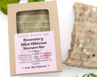 May include: A brown cardboard box with a white label that reads "Rosemary Mint Hibiscus Shampoo Bar 4 oz (approx.) Contains saponified oils of "coconut, olive, sunflower, castor & grapeseed. "cocoa butter. "hibiscus powder. rosemary & peppermint essential oils "organic *Katie Mae Naturals*"  A bar of soap is visible inside the box and a second bar of soap is sitting on a white surface.