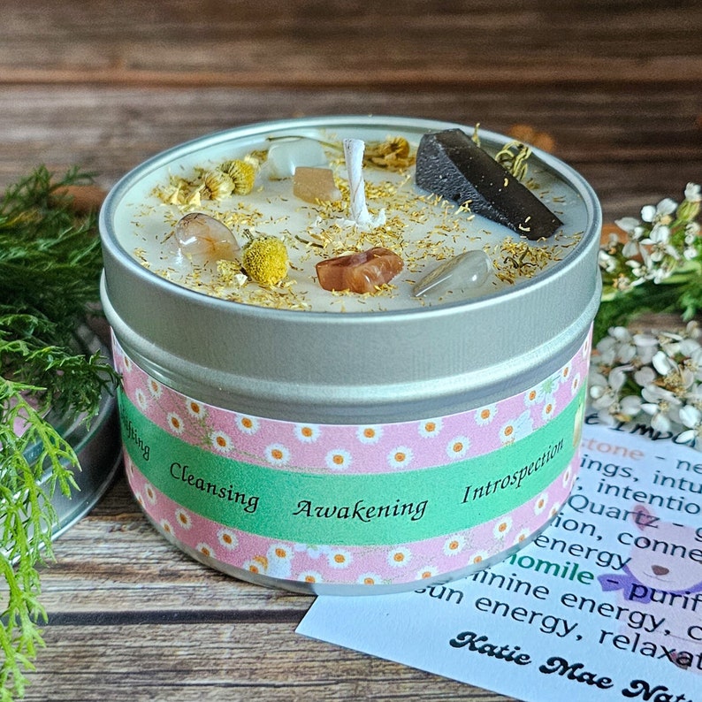 May include: A silver tin candle with a white wax top. The candle is decorated with small crystals, dried flowers, and gold glitter. The label on the tin reads "Cleansing Awakening Introspection".
