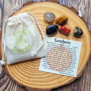 May include: A set of five crystals on a wooden surface with a small white bag and a card that reads "Samhain is 'summers end' - a time to celebrate Autumn, honor the dead, and rid ourselves of things not needed. Release old patterns that no longer serve us by writing them down and burn in a bonfire or cauldron. Celebrate the third harvest preparing a feast and set a place for loved ones lost. Carve a pumpkin with a spooky face to ward off evil and light a candle inside to welcome the kindred spirits. Useful herbs/foods: mugwort, sage, rosemary, and squash, apples, corn, nuts. Katie Mae Naturals.net"