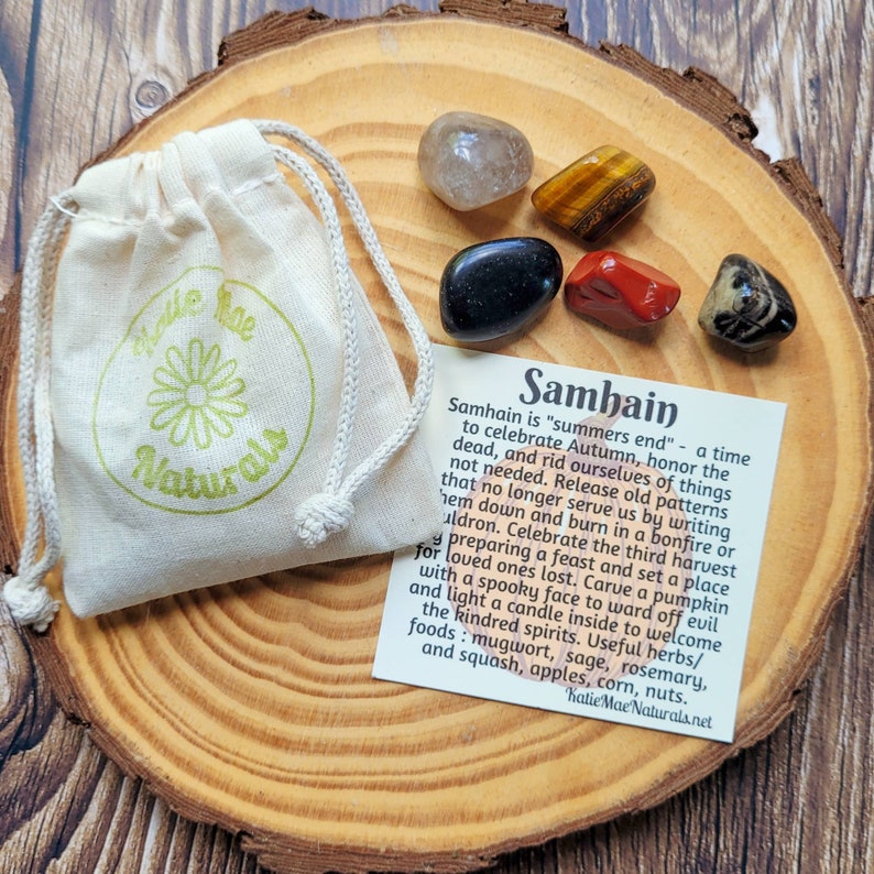 May include: A set of five crystals on a wooden surface with a small white bag and a card that reads "Samhain is 'summers end' - a time to celebrate Autumn, honor the dead, and rid ourselves of things not needed. Release old patterns that no longer serve us by writing them down and burn in a bonfire or cauldron. Celebrate the third harvest preparing a feast and set a place for loved ones lost. Carve a pumpkin with a spooky face to ward off evil and light a candle inside to welcome the kindred spirits. Useful herbs/foods: mugwort, sage, rosemary, and squash, apples, corn, nuts. Katie Mae Naturals.net"