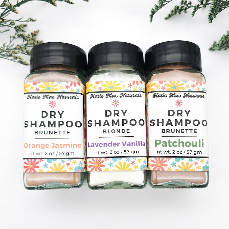May include: Three small glass jars with white lids containing dry shampoo for different hair colors. The jars are labeled "Katie Mae Naturals Dry Shampoo" with the hair color and scent listed below. The scents are Orange Jasmine, Lavender Vanilla, and Patchouli.